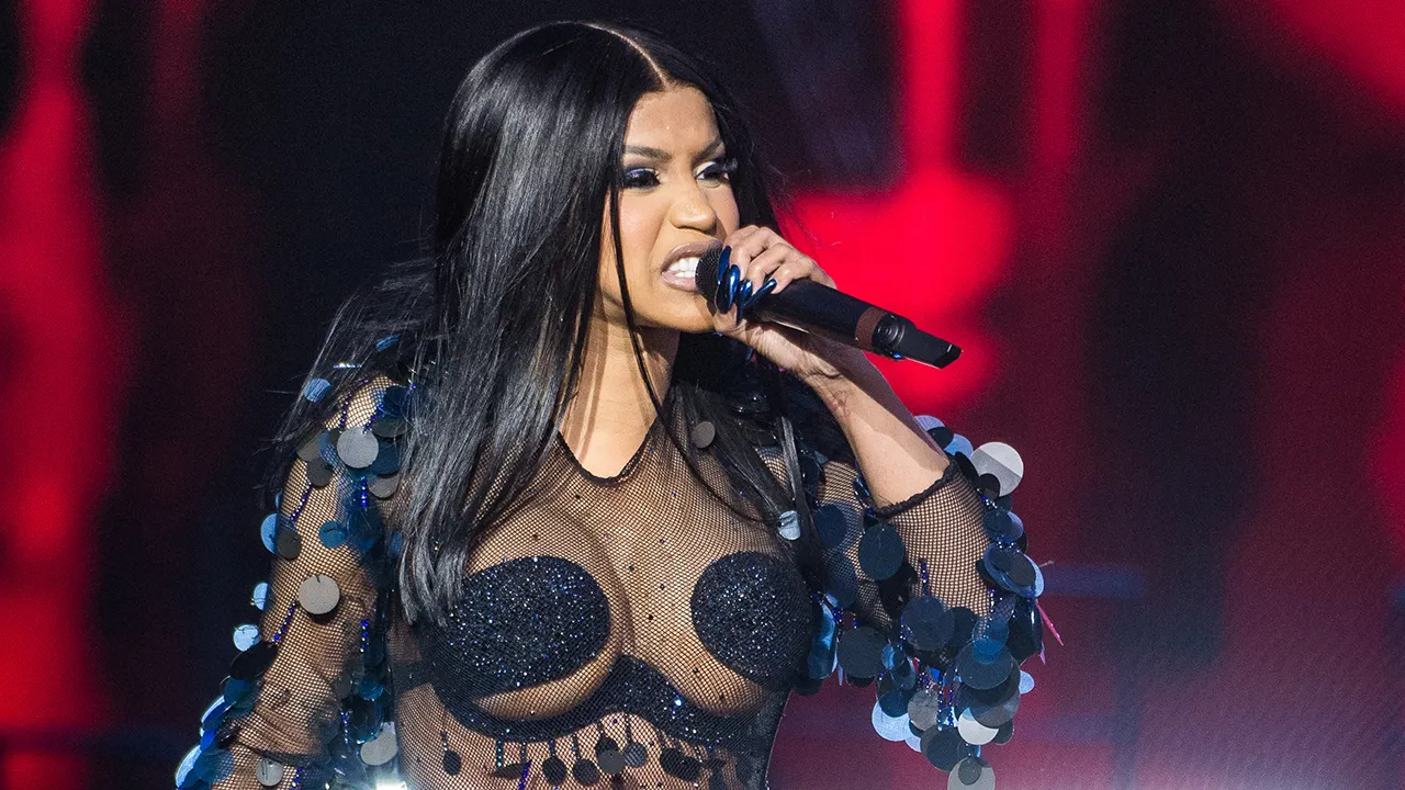 Cardi B wears a mesh black bodysuit on stage with large black circles around her chest singing into a microphone